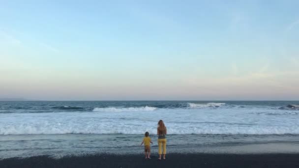 Genç bir kadın ve küçük oğlu okyanus kıyısında siyah volkanik kumla. Telefonla vurulmuş. — Stok video