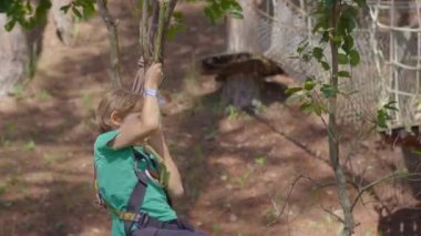 Macera parkında küçük bir çocuk. Emniyet kemeri takıyor. Yüksek bir ip patikasına tırmanıyor. Açık hava eğlence merkezi. Tırmanma faaliyetleri, kablo hatları ve her türlü engelden oluşuyor.
