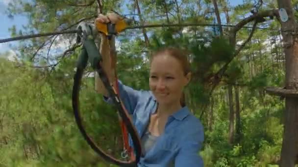 Een jonge vrouw rijdt op een zip-line in een avonturenpark. Ze draagt een veiligheidsharnas. Outdoor amusement centrum met klimactiviteiten bestaande uit zip-lijnen en allerlei obstakels. Langzame video — Stockvideo