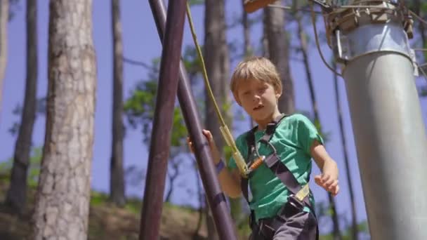 Egy kisfiú egy kalandparkban. Biztonsági övet visel. Egy magas kötélen mászik. Szabadtéri szórakoztató központ mászó tevékenységek álló vezetékek és mindenféle akadály — Stock videók