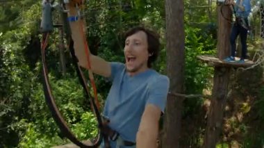 Genç bir adam macera parkında teleferik kullanıyor. Emniyet kemeri takıyor. Yüksek bir ip patikasına tırmanıyor. Açık hava eğlence merkezi. Tırmanma faaliyetleri kablo hatları ve her türlü şeyden oluşuyor.