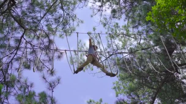 En ung man i en äventyrspark. Han har säkerhetssele. Han klättrar på ett rep. Utomhus nöjescentrum med klättring aktiviteter bestående av zip linjer och alla typer av hinder — Stockvideo