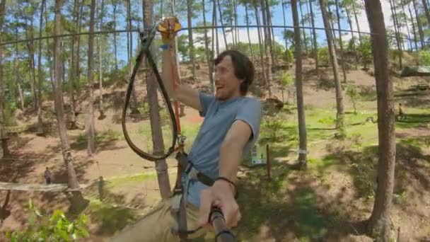 Ein junger Mann fährt in einem Abenteuerpark eine Seilrutsche. Er trägt einen Sicherheitsgurt. Er klettert auf einem Hochseilgarten. Outdoor-Vergnügungszentrum mit Kletteraktivitäten bestehend aus Seilrutschen und allerlei — Stockvideo