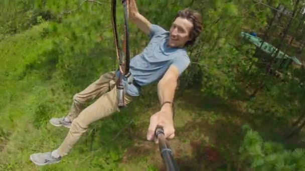 En ung man åker linbana i en äventyrspark. Han har säkerhetssele. Han klättrar på ett rep. Utomhus nöjescentrum med klättring verksamhet bestående av zip linjer och alla typer av — Stockvideo