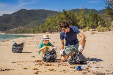 Plajı temizleyen eldivenli baba ve oğul kirli denizi temizleyen plastik torbaları topluyorlar. Çocukların doğal eğitimi. İnsan yapımı kumsalda dökülen çöplerle ilgili bir sorun.