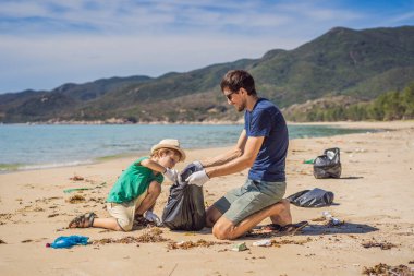 Plajı temizleyen eldivenli baba ve oğul kirli denizi temizleyen plastik torbaları topluyorlar. Çocukların doğal eğitimi. İnsan yapımı kumsalda dökülen çöplerle ilgili bir sorun.