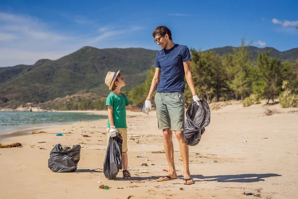 Vater und Sohn in Handschuhen säubern den Strand und sammeln Plastiktüten auf, die das Meer verschmutzen. Natürliche Erziehung der Kinder. Problem des verschütteten Mülls Müll am Strand Sand durch künstliche verursacht — Stockfoto