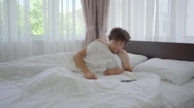 Genç bir adam mutlu bir şekilde yatakta uyanıyor ve telefonunu kontrol ediyor.