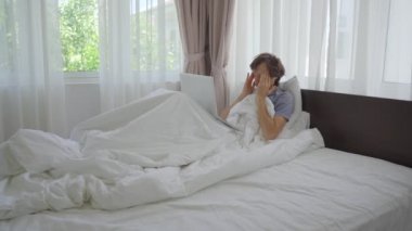 Yatakta üzgün genç bir adam video konferansında biriyle konuşur. Not defteri kullanarak. Sosyal izolasyon sırasında evde oturmaktan yoruldu.