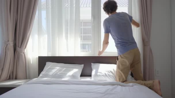 Un joven abre ventanas para que entre la luz de la mañana y se acuesta sonriendo en la cama — Vídeo de stock