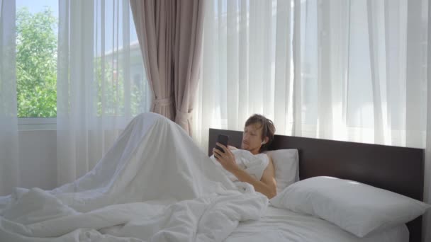 A young man in bed is talking to someone using a smartphone and making notes in a copybook — Stock Video