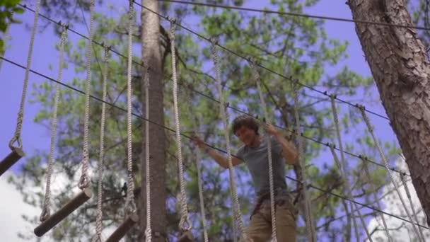 En ung man i en äventyrspark. Han har säkerhetssele. Han klättrar på ett rep. Utomhus nöjescentrum med klättring aktiviteter bestående av zip linjer och alla typer av hinder — Stockvideo