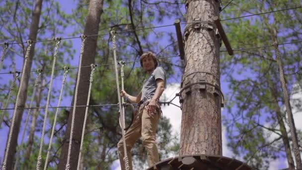 一个年轻人在一个探险公园里他戴着安全带.他爬上一条高绳的小径.户外娱乐中心，由拉链和各种障碍物组成的攀爬活动 — 图库视频影像