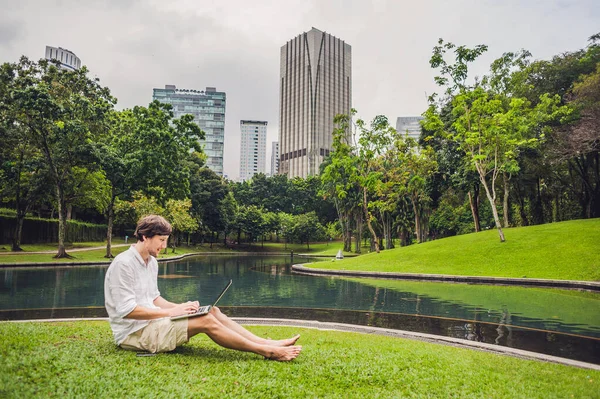Mann Geschäftsmann oder Student in Freizeitkleidung mit Laptop in einem tropischen Park vor dem Hintergrund von Wolkenkratzern. Bekleidet mit einem weißen Hemd, beigen Shorts. Mobiles Bürokonzept — Stockfoto