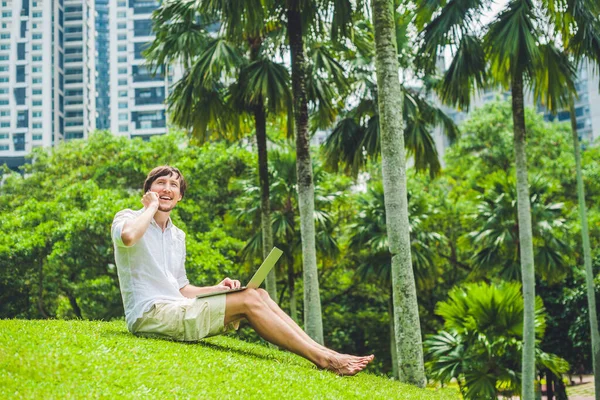 Mann Geschäftsmann oder Student in Freizeitkleidung mit Laptop in einem tropischen Park vor dem Hintergrund von Wolkenkratzern. Bekleidet mit einem weißen Hemd, beigen Shorts. Mobiles Bürokonzept — Stockfoto