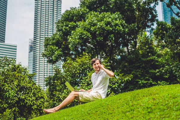 高層ビルの背景にある熱帯公園でラップトップを使用してカジュアルな服装のビジネスマンや学生。白いシャツを着て、ベージュのショートパンツ。モバイルオフィスのコンセプト — ストック写真