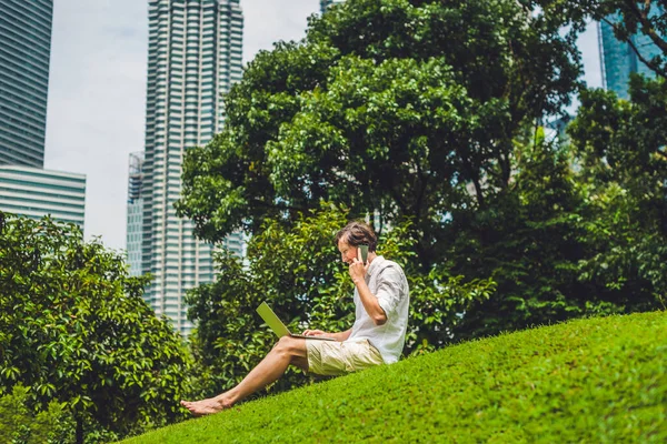 高層ビルの背景にある熱帯公園でラップトップを使用してカジュアルな服装のビジネスマンや学生。白いシャツを着て、ベージュのショートパンツ。モバイルオフィスのコンセプト — ストック写真