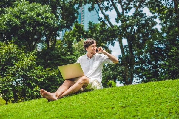 Чоловік-підприємець або студент у повсякденному одязі, використовуючи ноутбук у тропічному парку на тлі хмарочосів. Сукня в білій сорочці, бежеві шорти. Концепція мобільного офісу — стокове фото