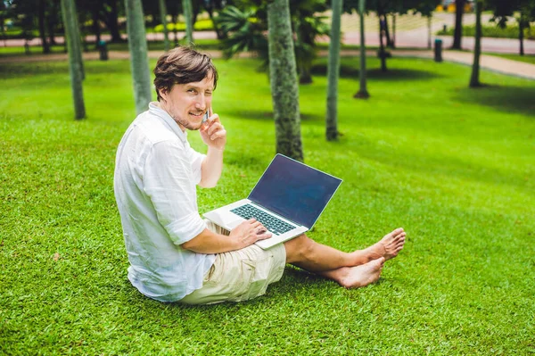 Чоловік-підприємець або студент у повсякденному одязі, використовуючи ноутбук у тропічному парку на тлі хмарочосів. Сукня в білій сорочці, бежеві шорти. Концепція мобільного офісу — стокове фото