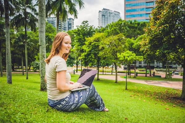 En ung kvinna i ledig klänning med bärbar dator i en tropisk park på bakgrunden av skyskrapor. Mobil Office koncept — Stockfoto