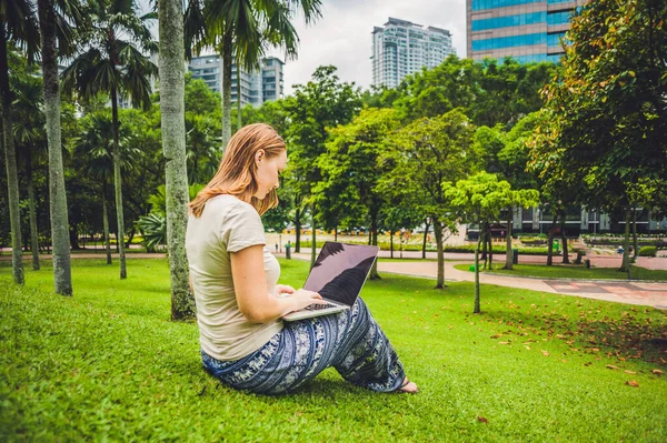 Молода жінка в повсякденному одязі використовує ноутбук у тропічному парку на тлі хмарочосів. Концепція мобільного офісу — стокове фото