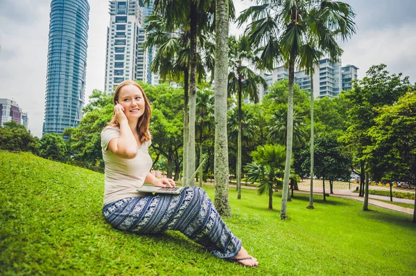 Молода жінка в повсякденному одязі використовує ноутбук у тропічному парку на тлі хмарочосів. Концепція мобільного офісу — стокове фото