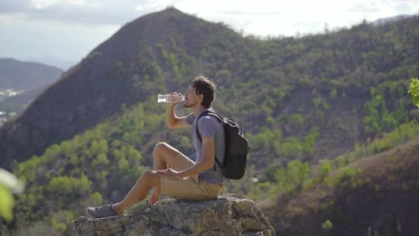 Młody człowiek siedzi na skale w górach i pije wodę. Pojęcie turystyki pieszej — Wideo stockowe