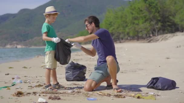 Un hombre y un niño con guantes recogen basura plástica en una playa. El problema de la basura en la arena de la playa causada por la contaminación causada por el hombre. Eco campañas para limpiar el medio ambiente. Voluntariado ecológico — Vídeo de stock