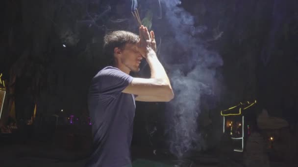 Um jovem europeu está rezando segurando varas de aroma de fumo em suas mãos. Ele está no maior templo caverna nas montanhas de mármore um complexo de templos budistas, um destino turístico famoso no — Vídeo de Stock