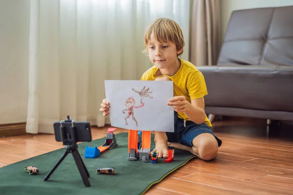 Zůstávám ve spojení. Šťastný mladý chlapec se dívá na svůj mobilní telefon a usmívá se při hraní s hračkami. Chlapec ukáže svou kresbu někomu prostřednictvím videokomunikace během vlastní izolace kvůli karanténě — Stock fotografie