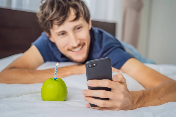 Quarantine birthday. Man with a smartphone and birthday cake-apple with candle. Stay home while quarantine. Online call. Video conference