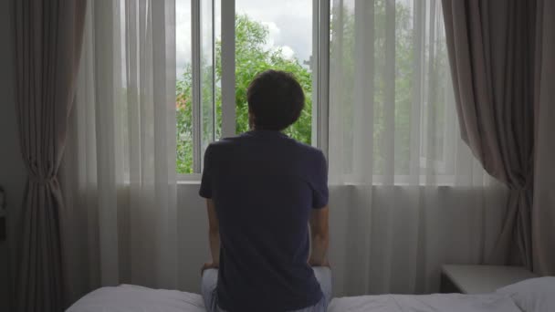 A young man in a mandatory quarantine. He sits and looks outside the window. He stays in one room for two weeks. He did not shave for long time, he is stressed and upset. Staying in quarantine concept — Stock Video