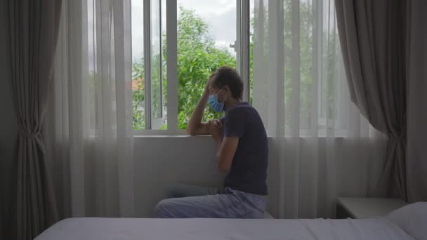 A young man in a mandatory quarantine. He sits and looks outside the window. He stays in one room for two weeks. He did not shave for long time, he is stressed and upset. Staying in quarantine concept — Stock Video