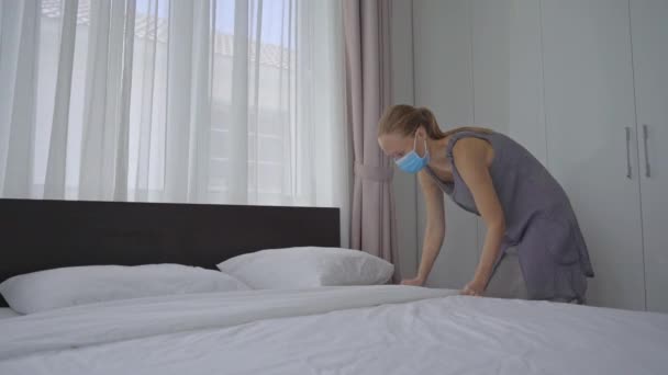 Uma jovem mulher em uma máscara facial médica muda de roupa de cama em um quarto de hotel. O hotel funciona como uma instalação de quarentena especial para pessoas que chegam do exterior. Viagem durante o conceito COVID — Vídeo de Stock