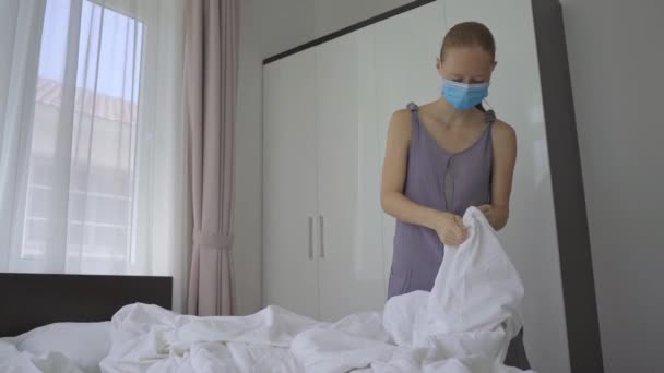 Seorang wanita muda bertopeng wajah medis mengubah linen tidur di kamar hotel. Hotel ini berfungsi sebagai fasilitas karantina khusus untuk orang-orang yang datang dari luar negeri. Perjalanan selama konsep COVID — Stok Video