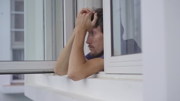 A young man in mandatory quarantine. He stays in one room for two weeks. He did not shave for a long time, he is stressed and upset. He sits by the window and looks outside. Staying in quarantine — Stock Video