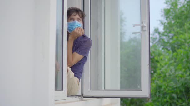 A young man in mandatory quarantine. He stays in one room for two weeks. He did not shave for a long time, he is stressed and upset. He sits by the window wearing a facemask and looks outside. Staying — Stock Video
