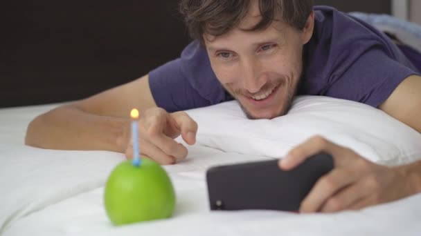 Ένας νεαρός σε υποχρεωτική καραντίνα. Γιορτάζει τα γενέθλιά του έχοντας ένα μήλο αντί για τούρτα. Μιλάει σε ένα φίλο ή συγγενή χρησιμοποιώντας smartphone. Έννοια του δεύτερου κύματος Covid-19 — Αρχείο Βίντεο