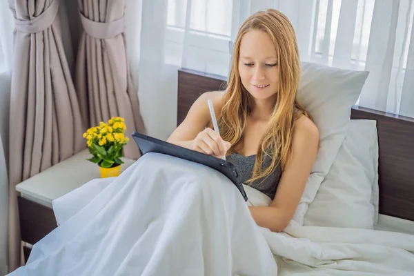 Woman draws on tablet with stylus in bed