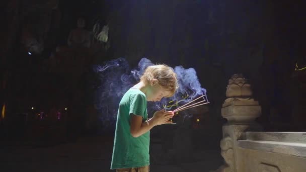 Tiro de câmera lenta de um menino que está orando segurando varas de aroma de fumaça em suas mãos. Ele está no maior templo caverna nas montanhas de mármore um complexo de templos budistas, um famoso turista — Vídeo de Stock