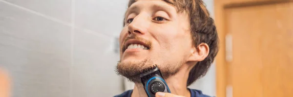 Handsome young bearded man trimming his beard with a trimmer BANNER, LONG FORMAT