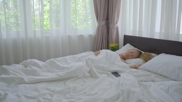 Молодая женщина в постели. Она не может проснуться, несмотря на звонок будильника на ее телефоне — стоковое видео
