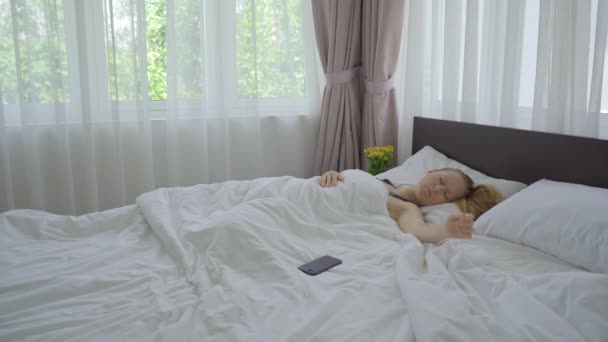 Een jonge vrouw in een bed. Ze kan niet wakker worden ondanks de wekker op haar telefoon. — Stockvideo