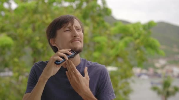 A young man shaves his beard standing on a balcony — Stock Video