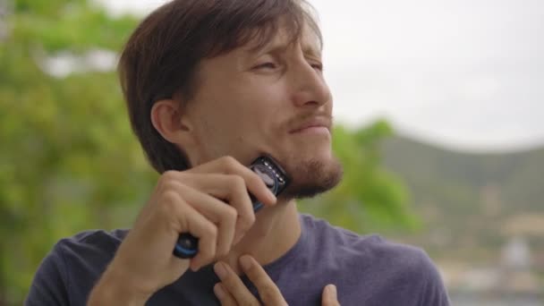 A young man shaves his beard standing on a balcony — Stock Video