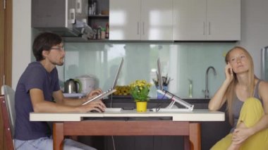 Arka planda mutfak olan genç bir kadın ve erkek oturma odalarında çalışıyorlar. İkisi de Covid-19 kısıtlamalarından dolayı uzaktan çalışmak zorunda. Kadın neşeyle telefonla konuşuyor.
