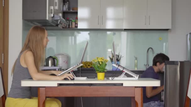 A young woman is working in the living room with a kitchen in the background. She has to work remotely due to the Covid-19 restrictions. The man is loudly searching for food in the refrigerator which — Stock Video