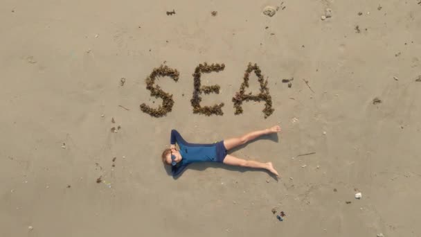 ภาพถ่ายทางอากาศของเด็กชายที่วางอยู่ข้างป้าย "ทะเล" ที่ทําจากสาหร่ายทะเลบนชายหาด หุ่นยนต์เคลื่อนตัวลง — วีดีโอสต็อก