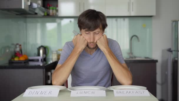 Un joven triste se sienta frente a una mesa con platos vacíos con signos de desayuno, almuerzo y cena. Concepto de ayuno de intervalo. Saltarse las comidas — Vídeos de Stock