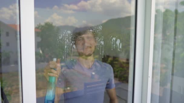 Молодий чоловік миє скляні двері, використовуючи рідину для чищення скла. Гендерна рівність — стокове відео